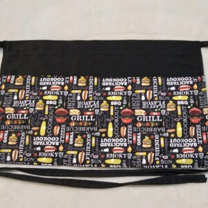 May include: A black waist apron featuring a repeating pattern of barbecue-themed graphics including burgers, hot dogs, corn, and grills, interspersed with text such as "GRILL," "SMOKY," "BACKYARD COOKOUT," and "FRIENDS" on a black background.
