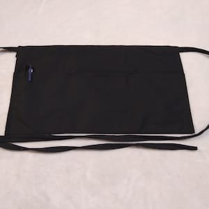 May include: A black apron with two long ties. The apron has a single pocket on the front with a blue pen sticking out.