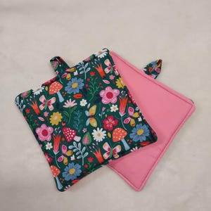 May include: Two square pot holders. One features a dark green floral pattern with mushrooms, butterflies, and flowers in pink, yellow, and blue. The other is solid pink. Both have a small loop for hanging.