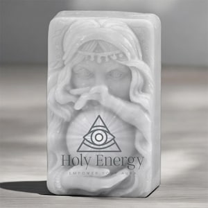 May include: A white rectangular candle featuring a carved relief of a woman's face, holding a crystal ball. The candle is inscribed with the words "Holy Energy" and "Empower Your Aura".