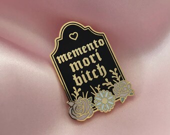 May include: A black enamel pin in the shape of a tombstone with the words "memento mori bitch" in gold lettering. The pin features a heart and floral accents in shades of pink, blue, and gold.