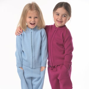 May include: Two young girls wearing matching knit sweaters and pants. The girl on the left is wearing a light blue sweater and pants, while the girl on the right is wearing a pink sweater and pants. Both girls are smiling.
