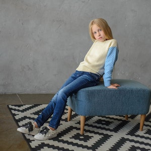 K&ouml;nnte beinhalten: Ein junges M&auml;dchen tr&auml;gt einen hellblauen und cremefarbenen Pullover, blaue Jeans und wei&szlig;e Turnschuhe und sitzt auf einem blauen, gepolsterten Hocker mit Holzbeinen. Der Hocker steht auf einem schwarz-wei&szlig;en Teppich.