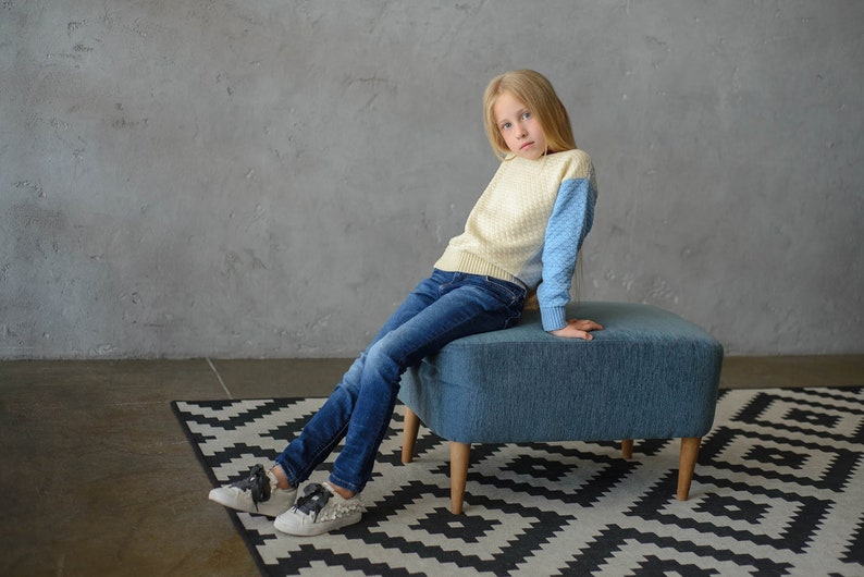 K&ouml;nnte beinhalten: Ein junges M&auml;dchen tr&auml;gt einen hellblauen und cremefarbenen Pullover, blaue Jeans und wei&szlig;e Turnschuhe und sitzt auf einem blauen, gepolsterten Hocker mit Holzbeinen. Der Hocker steht auf einem schwarz-wei&szlig;en Teppich.