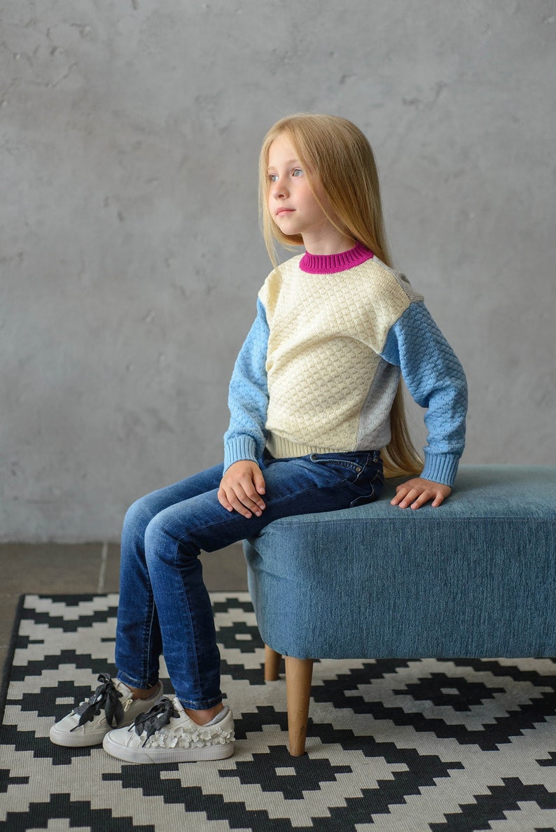 K&ouml;nnte beinhalten: Ein junges M&auml;dchen mit langen blonden Haaren sitzt auf einem blauen Ottoman und tr&auml;gt einen cremefarbenen, blauen und rosafarbenen Pullover und eine blaue Jeans. Sie tr&auml;gt wei&szlig;e Turnschuhe mit schwarzen Schn&uuml;rsenkeln und einer schwarzen Schleife. Der Ottoman steht auf einem schwarz-wei&szlig; gemusterten Teppich.