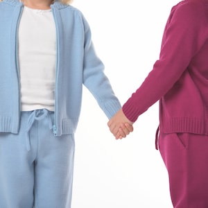 May include: Two children holding hands, one wearing a light blue knitted sweater and pants, the other wearing a burgundy knitted sweater and pants.