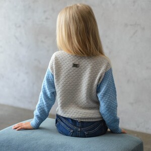 K&ouml;nnte beinhalten: Ein hellgrauer und blauer Strickpullover mit einem kleinen Metalletikett auf der R&uuml;ckseite. Der Pullover hat ein strukturiertes Muster und wird von einer Person getragen, die auf einem blauen Kissen sitzt.