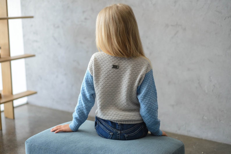 K&ouml;nnte beinhalten: Ein hellgrauer und blauer Strickpullover mit einem kleinen Metalletikett auf der R&uuml;ckseite. Der Pullover hat ein strukturiertes Muster und wird von einer Person getragen, die auf einem blauen Kissen sitzt.