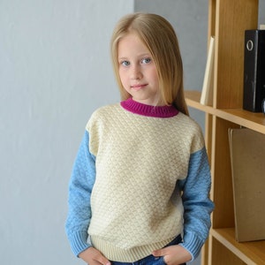 K&ouml;nnte beinhalten: Ein junges M&auml;dchen tr&auml;gt einen cremefarbenen, blauen und rosafarbenen Pullover mit einem strukturierten Muster. Sie tr&auml;gt eine blaue Jeans und hat die H&auml;nde in den Taschen. Sie steht vor einem B&uuml;cherregal aus Holz.
