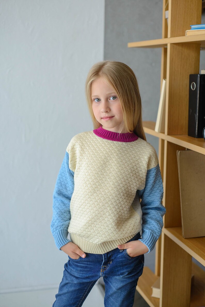 K&ouml;nnte beinhalten: Ein junges M&auml;dchen tr&auml;gt einen cremefarbenen, blauen und rosafarbenen Pullover mit einem strukturierten Muster. Sie tr&auml;gt eine blaue Jeans und hat die H&auml;nde in den Taschen. Sie steht vor einem B&uuml;cherregal aus Holz.