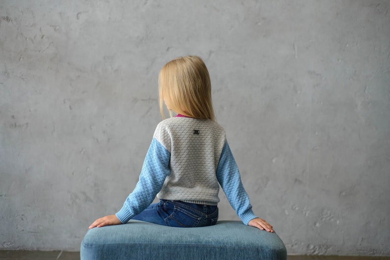 K&ouml;nnte beinhalten: Eine junge Person tr&auml;gt einen blau-grauen Pullover mit einem kleinen Logo auf dem R&uuml;cken und sitzt auf einer blauen gepolsterten Bank.