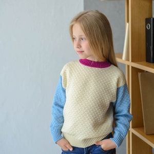 K&ouml;nnte beinhalten: Ein junges M&auml;dchen tr&auml;gt einen hellblauen und cremefarbenen Pullover mit einem rosa Saum am Hals. Der Pullover hat ein strukturiertes Muster. Das M&auml;dchen tr&auml;gt eine blaue Jeans und hat die H&auml;nde in den Taschen. Das M&auml;dchen steht vor einem B&uuml;cherregal aus Holz.