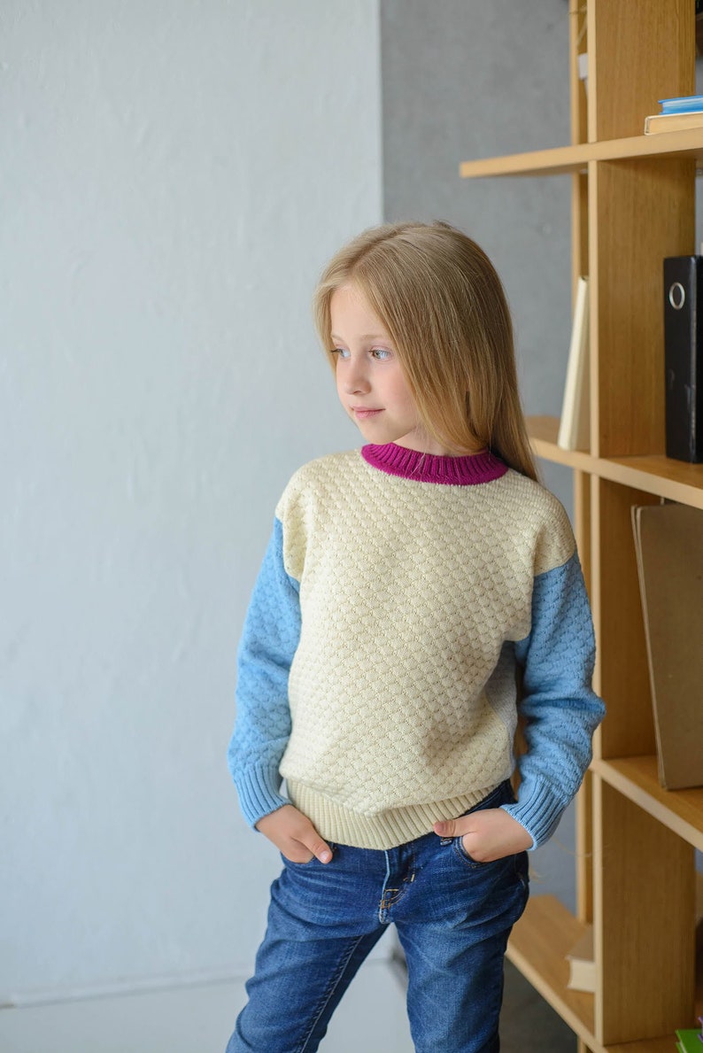 K&ouml;nnte beinhalten: Ein junges M&auml;dchen tr&auml;gt einen hellblauen und cremefarbenen Pullover mit einem rosa Saum am Hals. Der Pullover hat ein strukturiertes Muster. Das M&auml;dchen tr&auml;gt eine blaue Jeans und hat die H&auml;nde in den Taschen. Das M&auml;dchen steht vor einem B&uuml;cherregal aus Holz.