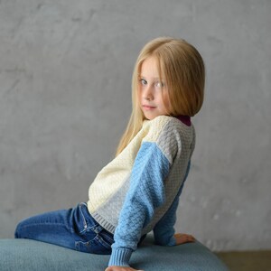 K&ouml;nnte beinhalten: Ein junges M&auml;dchen mit langen blonden Haaren sitzt auf einem blauen Hocker. Sie tr&auml;gt einen blau-wei&szlig;-grauen Pullover und eine blaue Jeans. Sie schaut &uuml;ber ihre Schulter in die Kamera.