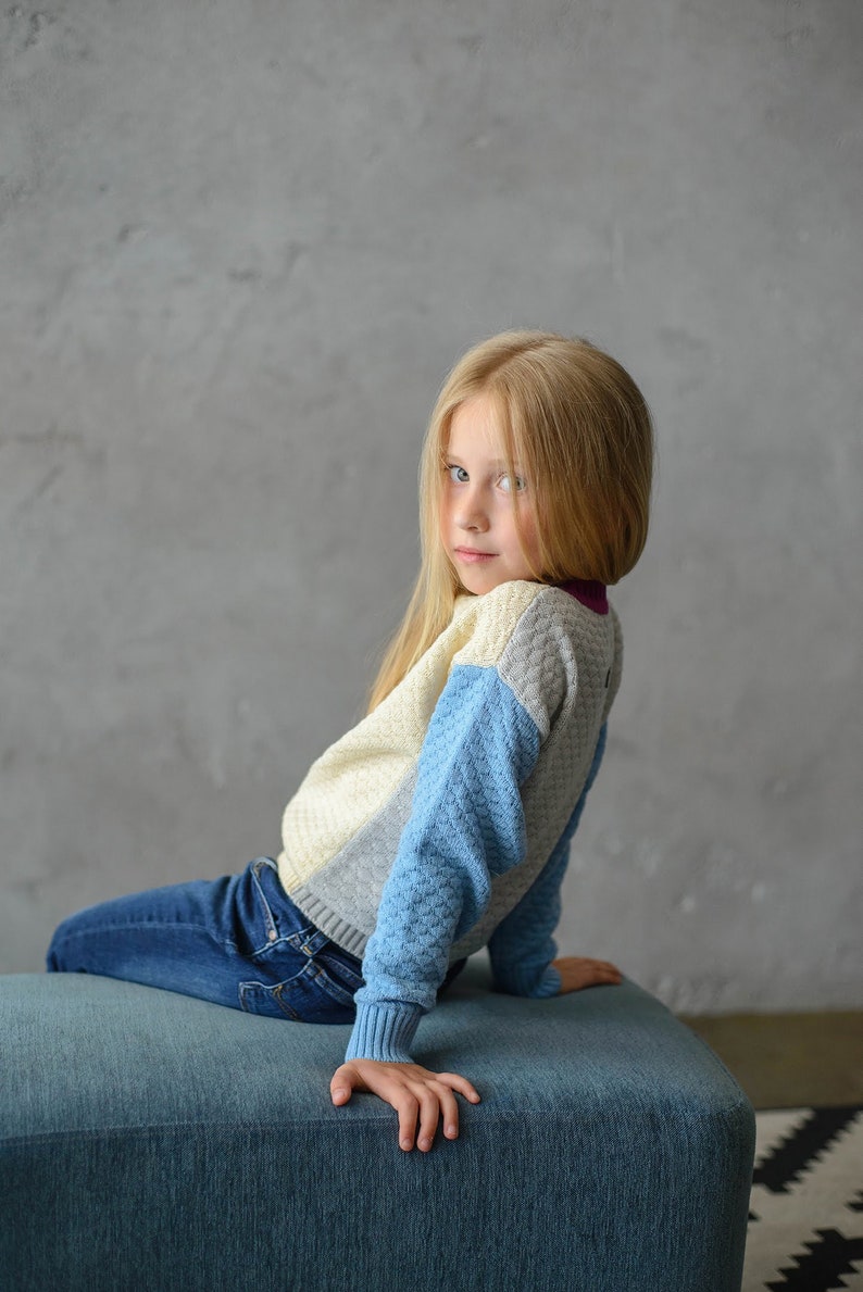 K&ouml;nnte beinhalten: Ein junges M&auml;dchen mit langen blonden Haaren sitzt auf einem blauen Hocker. Sie tr&auml;gt einen blau-wei&szlig;-grauen Pullover und eine blaue Jeans. Sie schaut &uuml;ber ihre Schulter in die Kamera.