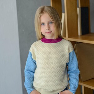 K&ouml;nnte beinhalten: Ein junges M&auml;dchen tr&auml;gt einen hellblauen und cremefarbenen Pullover mit rosa Kragen und blaue Jeans. Der Pullover hat ein strukturiertes Muster. Das M&auml;dchen steht vor einem B&uuml;cherregal aus Holz.