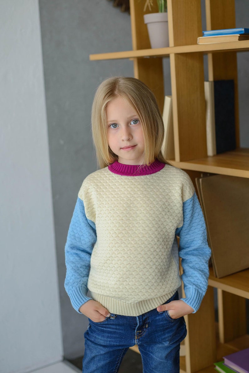 K&ouml;nnte beinhalten: Ein junges M&auml;dchen tr&auml;gt einen hellblauen und cremefarbenen Pullover mit rosa Kragen und blaue Jeans. Der Pullover hat ein strukturiertes Muster. Das M&auml;dchen steht vor einem B&uuml;cherregal aus Holz.