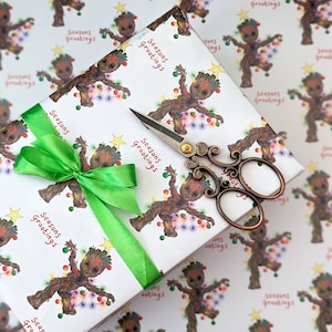 Puede incluir: Un regalo envuelto con una cinta y un lazo verdes, con papel estampado con un personaje de dibujos animados sosteniendo luces navideñas. El papel también tiene el texto "Seasons Greetings" en rojo. Unas tijeras decorativas descansan sobre el regalo.