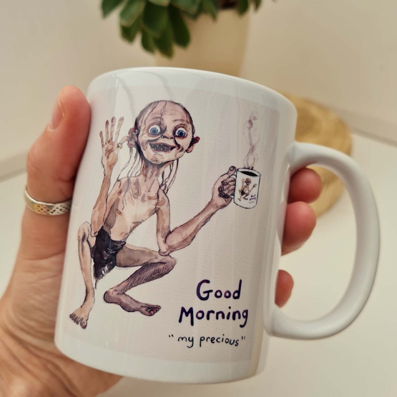 May include: White ceramic mug featuring a watercolor illustration of Gollum from The Lord of the Rings, holding a coffee cup. The mug has the text "Good Morning" and "my precious".