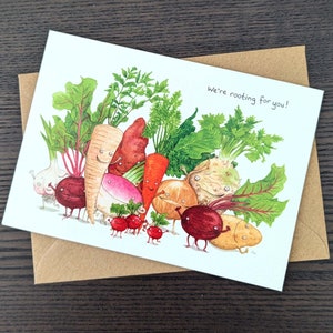 May include: A greeting card featuring a watercolour illustration of various vegetables with faces. The vegetables are arranged in a circle and the text "We're rooting for you!" is written in black at the top of the card.