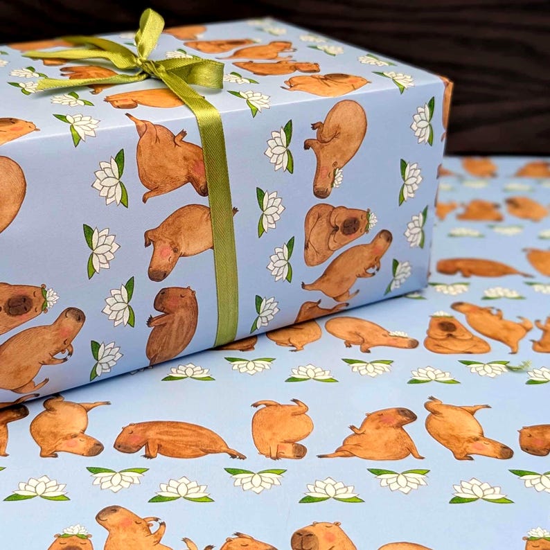 May include: A wrapped gift box featuring light blue wrapping paper with a repeating pattern of brown capybaras and white lotus flowers. The box is tied with a green ribbon. The wrapping paper is also visible in the background.