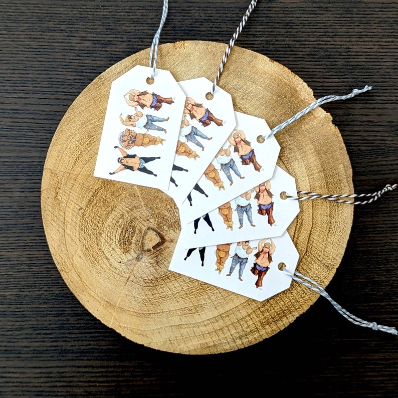May include: A collection of white gift tags with illustrations of a person in various poses, wearing cowboy attire. Each tag is strung with a gray and white twine. The tags are arranged on a wooden slice, set against a dark wood background.