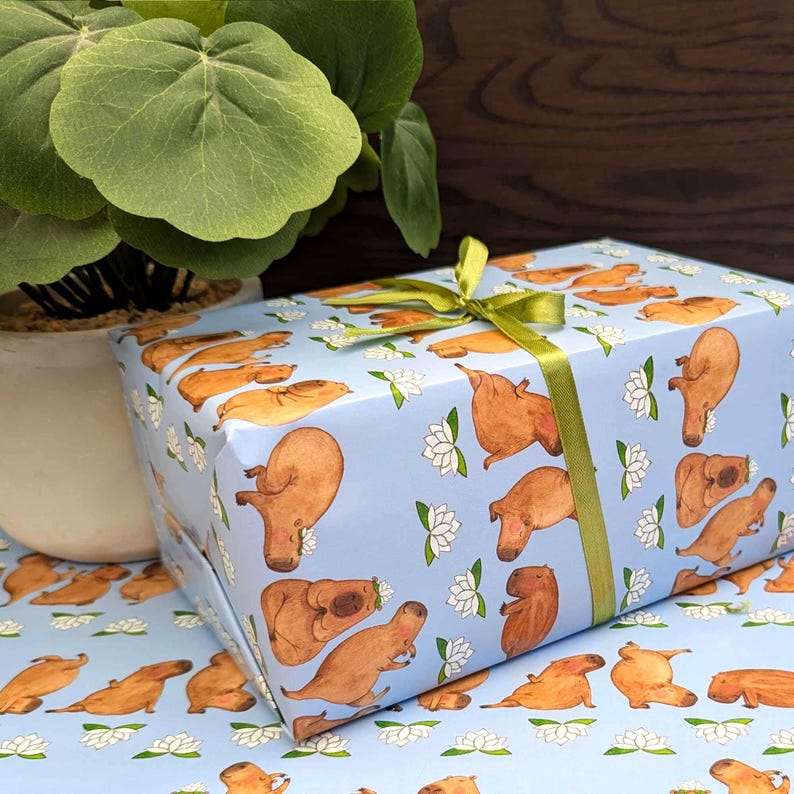 May include: A wrapped gift box featuring a light blue background with a repeating pattern of brown capybaras and white lotus flowers. The box is tied with a green ribbon and bow. A potted plant is in the background.