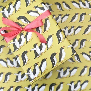 Puede incluir: Papel de regalo amarillo con un patr&oacute;n repetitivo de ping&uuml;inos negros y blancos. Los ping&uuml;inos est&aacute;n en varias poses, algunos est&aacute;n de pie, algunos est&aacute;n nadando y algunos est&aacute;n volando. El papel est&aacute; decorado con una cinta roja.