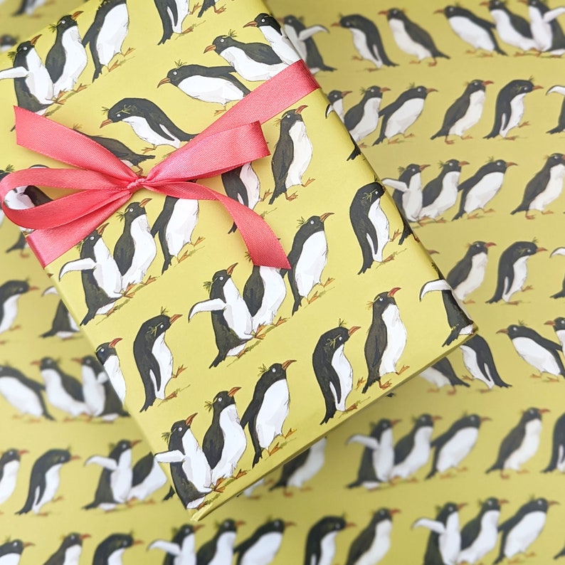 Puede incluir: Papel de regalo amarillo con un patr&oacute;n repetitivo de ping&uuml;inos negros y blancos. Los ping&uuml;inos est&aacute;n en varias poses, algunos est&aacute;n de pie, algunos est&aacute;n nadando y algunos est&aacute;n volando. El papel est&aacute; decorado con una cinta roja.
