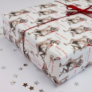 Könnte beinhalten: Ein Geschenk, verpackt in weißem Papier mit einer sich wiederholenden Illustration einer Kreatur mit Weihnachtsmütze. Die Worte "Happy Christmas" sind rot gedruckt. Das Geschenk ist mit rotem Garn und einer roten Schleife gebunden.