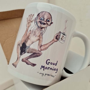May include: White ceramic mug featuring a watercolor illustration of a character holding a smaller mug with steam rising. The mug has the text "Good Morning" and "my precious" in purple script.