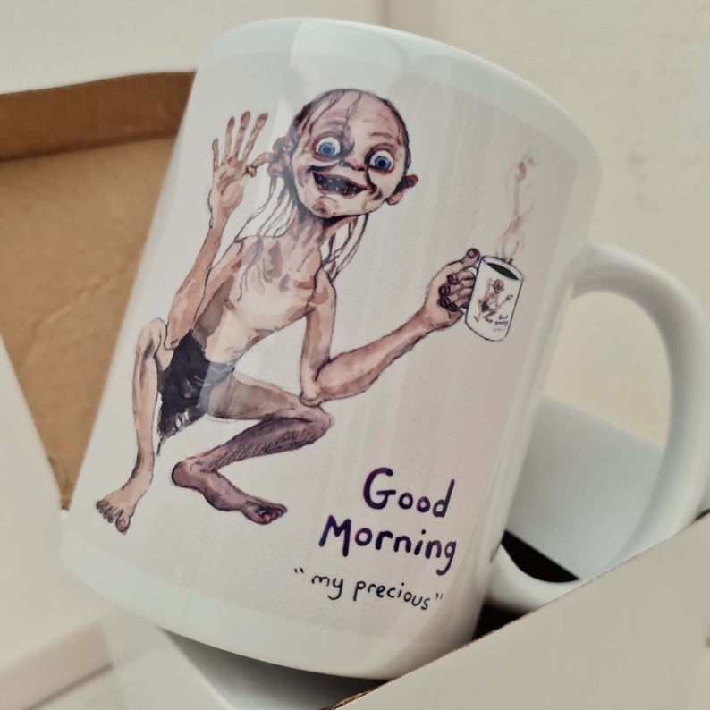 May include: White ceramic mug featuring a watercolor illustration of a character holding a smaller mug with steam rising. The mug has the text "Good Morning" and "my precious" in purple script.
