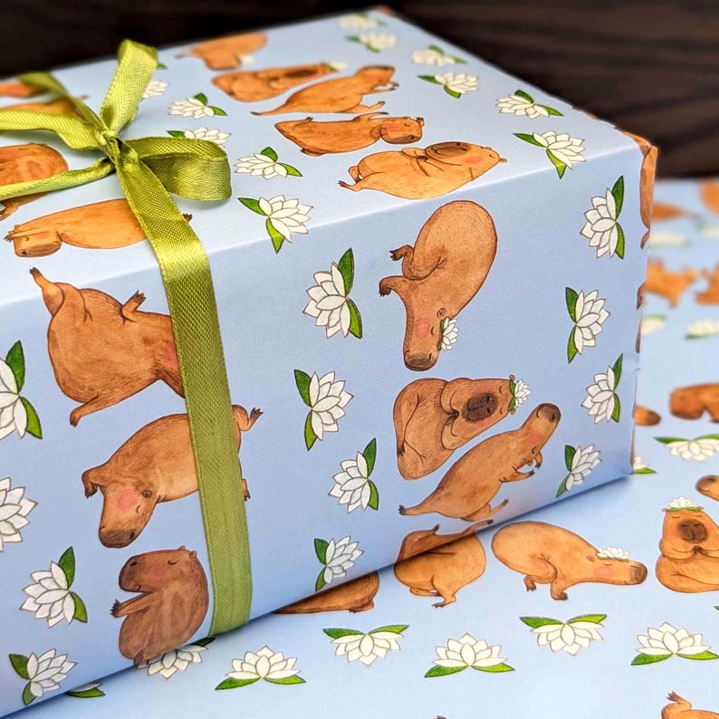 May include: A wrapped gift box featuring light blue wrapping paper with a pattern of brown capybaras and white lotus flowers. A green ribbon is tied around the box.