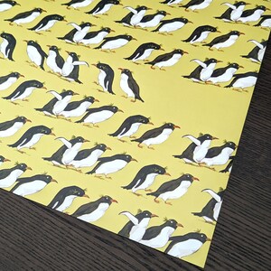 Puede incluir: Un papel de regalo amarillo con un patr&oacute;n repetitivo de ping&uuml;inos negros y blancos. Los ping&uuml;inos miran en diferentes direcciones y algunos est&aacute;n de pie mientras que otros nadan.