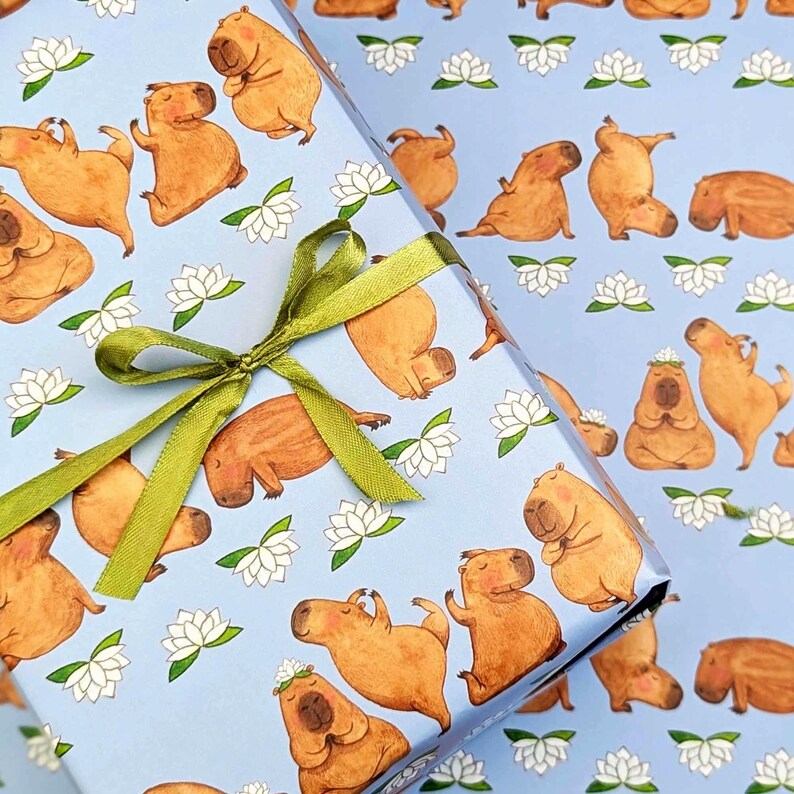 May include: Light blue wrapping paper featuring a repeating pattern of brown capybaras in various yoga poses and white lotus flowers. A green ribbon is tied in a bow around the wrapped paper, adding a decorative touch.