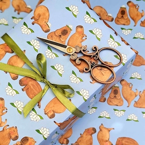 May include: A gift box wrapped in light blue paper featuring capybara illustrations and white lotus flowers. A green ribbon and decorative wooden scissors are on top. The scissors have a gold-colored rivet.