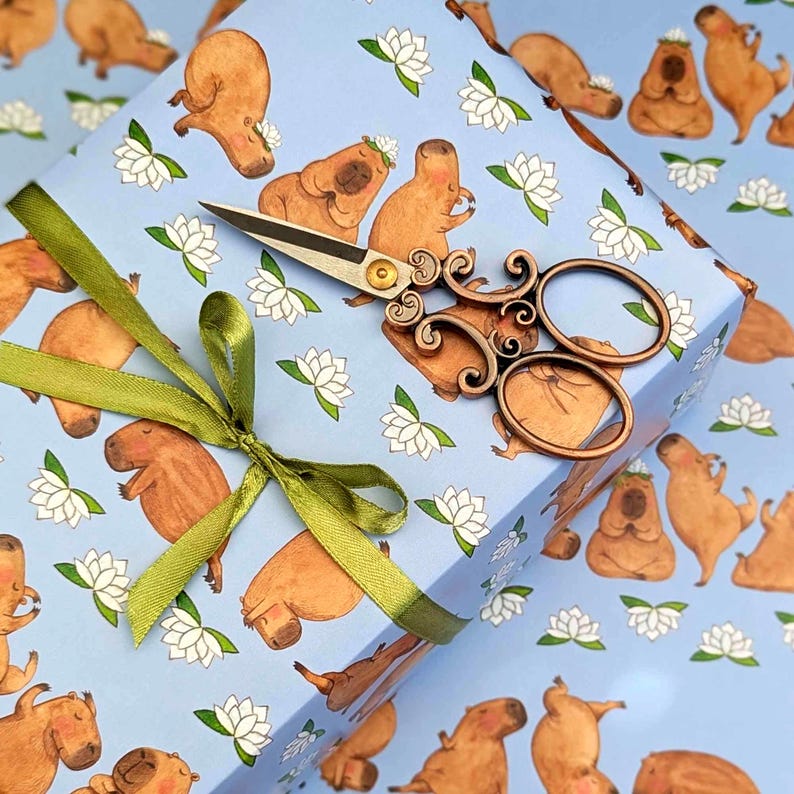 May include: A gift box wrapped in light blue paper featuring capybara illustrations and white lotus flowers. A green ribbon and decorative wooden scissors are on top. The scissors have a gold-colored rivet.