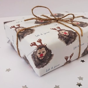May include: White gift wrap featuring a repeating pattern of a man with reindeer antlers and a red nose. The words "Let it Snow" are written on the wrap. The gift is tied with brown twine.
