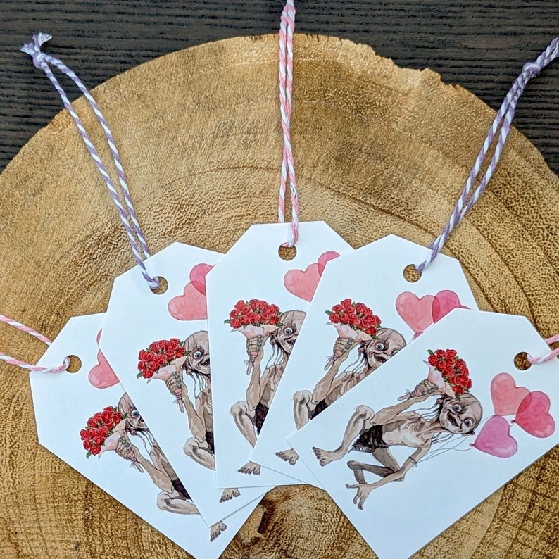 May include: Gift tags featuring a watercolour illustration of a creature holding a bouquet of red roses and pink heart-shaped balloons. The tags have a white background and are strung with pink and purple twine.