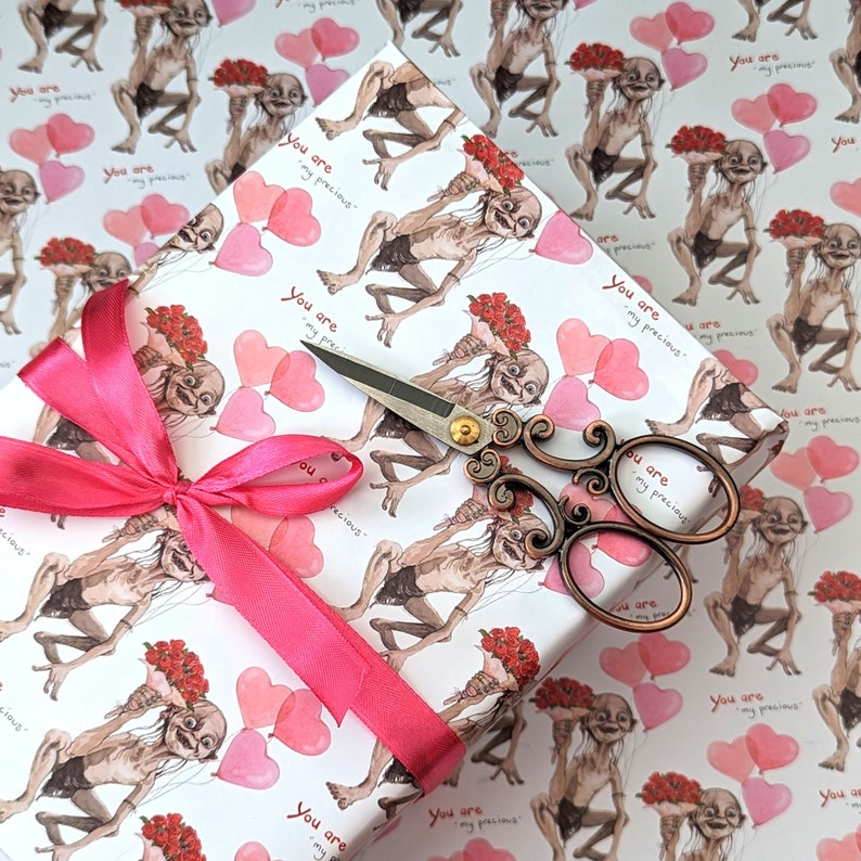 May include: Gift wrap with a repeating pattern of a creature holding flowers and pink heart balloons, with the text "You are my precious." A pink ribbon and decorative scissors are on top of the wrapped gift.