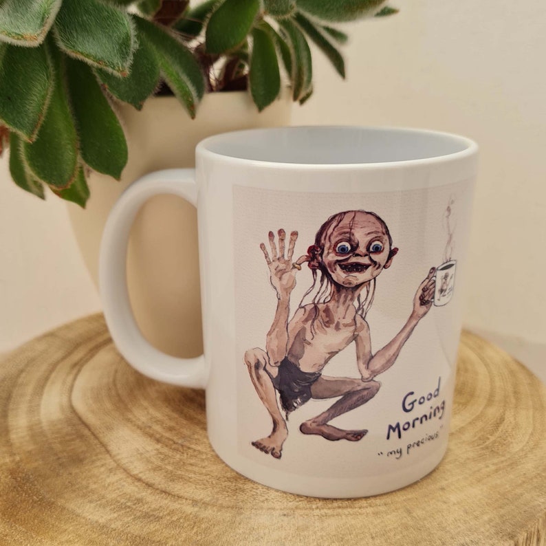 May include: White ceramic mug featuring a watercolor illustration of a character holding a coffee cup, with the text "Good Morning, my precious". The mug has a curved handle and is set against a wooden surface.