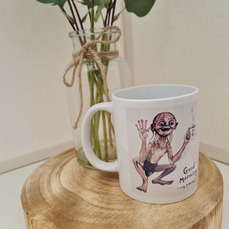 May include: White ceramic mug featuring a cartoon illustration of Gollum from The Lord of the Rings, holding a cup of coffee. The mug has the text "Good Morning... my precious" printed on it. The mug is on a wooden surface.