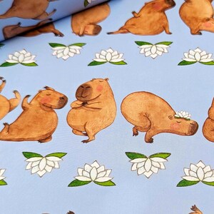 May include: Light blue fabric featuring a repeating pattern of cartoon capybaras in yoga poses, interspersed with white lotus flowers with green leaves. The capybaras are various shades of brown and tan.