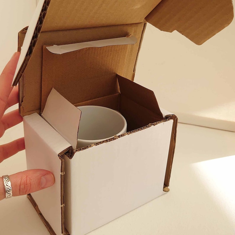 May include: A white ceramic mug inside a white cardboard box with a brown cardboard insert. The box is partially open, revealing the mug. The box is being held by a hand with a silver ring.