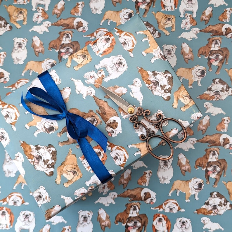 May include: A sheet of wrapping paper with a repeating pattern of brown and white bulldogs. The paper is decorated with a blue ribbon and a pair of silver scissors.