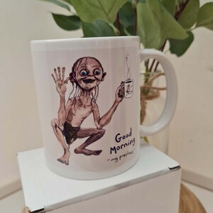 May include: White ceramic mug featuring a cartoon illustration of Gollum from The Lord of the Rings, holding a coffee cup. The mug has the text "Good Morning" and "my precious".