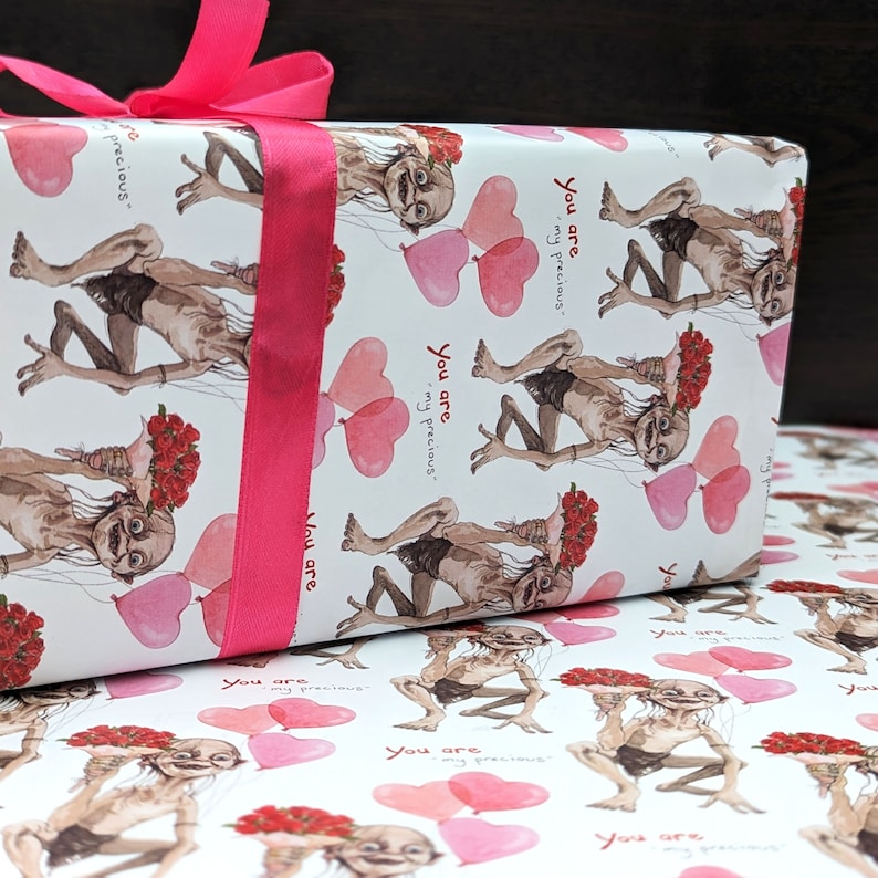 May include: Gift-wrapped present with white paper featuring a repeating pattern of a character holding flowers and heart-shaped balloons. The text "You are my precious" is also printed on the paper. A bright pink ribbon is tied around the gift.