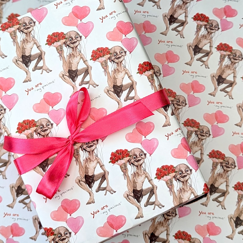 May include: Wrapping paper featuring a repeating pattern of a cartoon character holding flowers and heart-shaped balloons. The text "You are my precious" is printed on the paper. A pink ribbon is tied in a bow on top.
