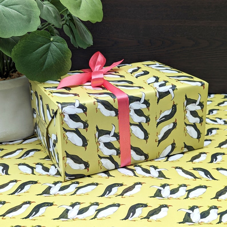 Puede incluir: Una caja de regalo amarilla con una cinta rosa y un patr&oacute;n de ping&uuml;ino. La caja est&aacute; envuelta en papel de regalo con tem&aacute;tica de ping&uuml;inos.