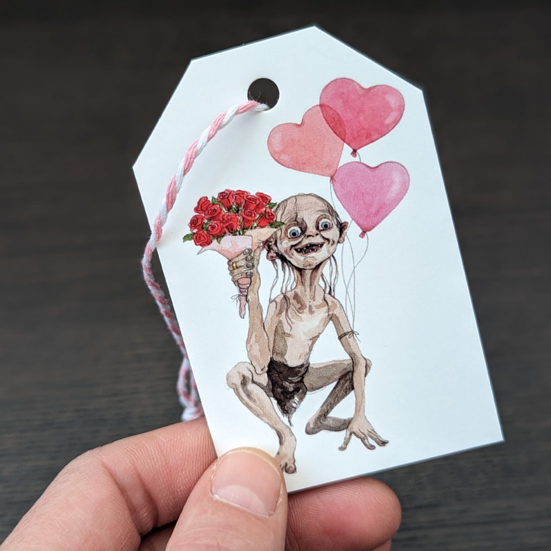 May include: A white gift tag featuring a watercolour illustration of a character holding a bouquet of red roses and pink heart-shaped balloons. The tag has a hole at the top with pink and white twine. The character is from a fantasy novel.